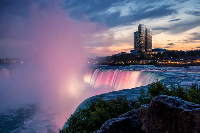 Editorial photograph of Niagara Falls at dusk with a modern glass casino resort tower in the background, warm evening lights, colorful mist rising from the falls